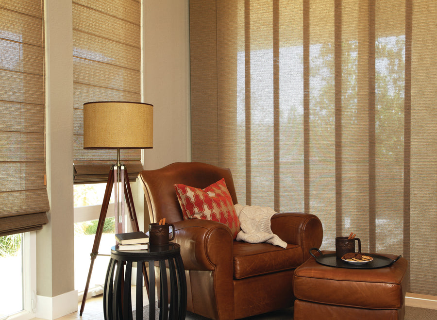 A cozy corner features a brown leather armchair, patterned pillows, matching ottoman, round side table, tripod floor lamp, and large windows with Skyline® Gliding Window Panels for the perfect natural light window treatment.