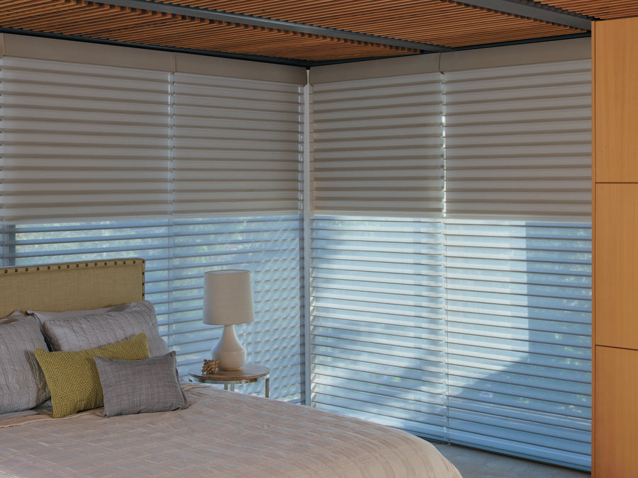 A modern bedroom features a light bed, pillows, bedside table with a white lamp, and large windows adorned with Nantucket™ Window Shadings, whose fabric vanes gently diffuse light. A wooden ceiling and cabinet bring warmth to the space.