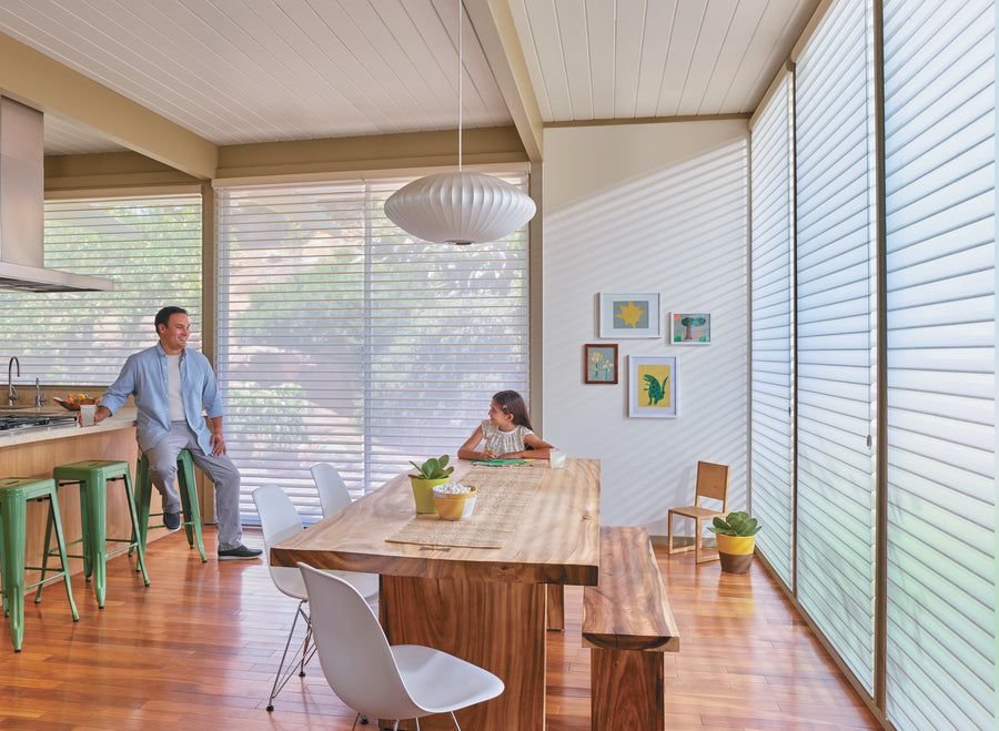 A man and a girl sit in a bright, modern dining room with wooden furniture and large windows featuring Nantucket™ Window Shadings. The fabric vanes softly diffuse sunlight, casting gentle shadows on the floor and framed art along the white wall.