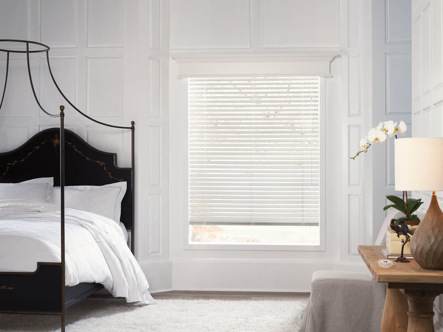 A bright bedroom with white walls, a dark four-poster bed with white bedding, and a large window featuring Parkland® Wood Blinds. A wooden desk with lamp, white orchid, and beige chair sits on a white rug.