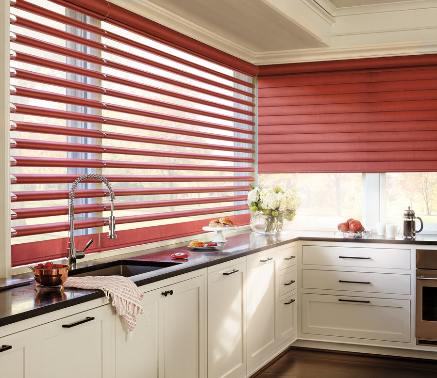 A bright kitchen with white cabinets, dark countertops, and large corner windows features Pirouette® Window Shadings in red for light filtration and UV protection. Sunlight streams in, illuminating flowers, fruit, and dishes on the counters.