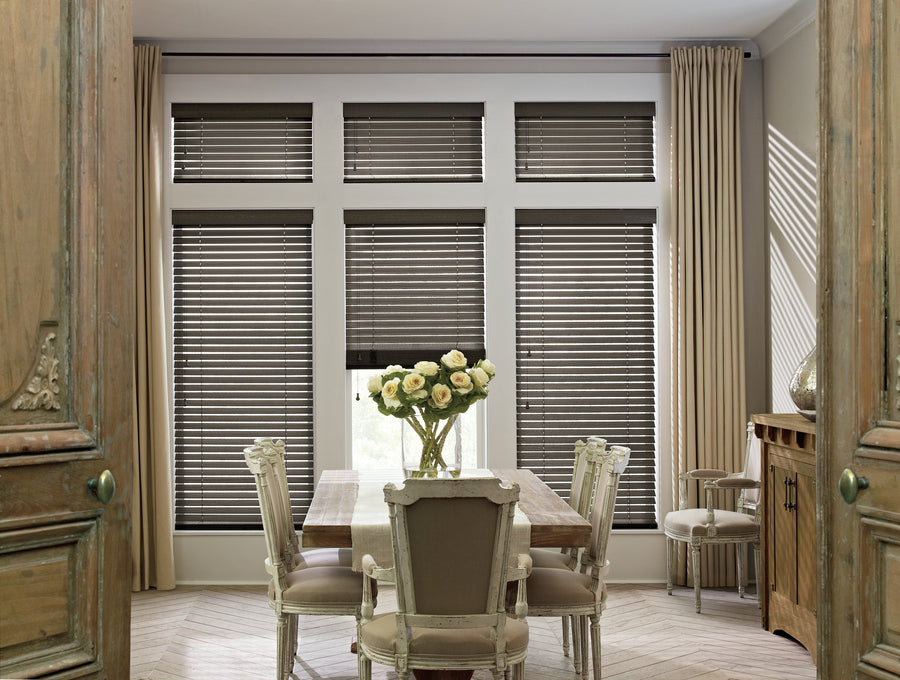 A dining room with a wooden table, six elegant chairs, white flowers in a vase, large windows with Parkland® Wood Blinds featuring hardwood slats, floor-length beige curtains, and light wood herringbone flooring.