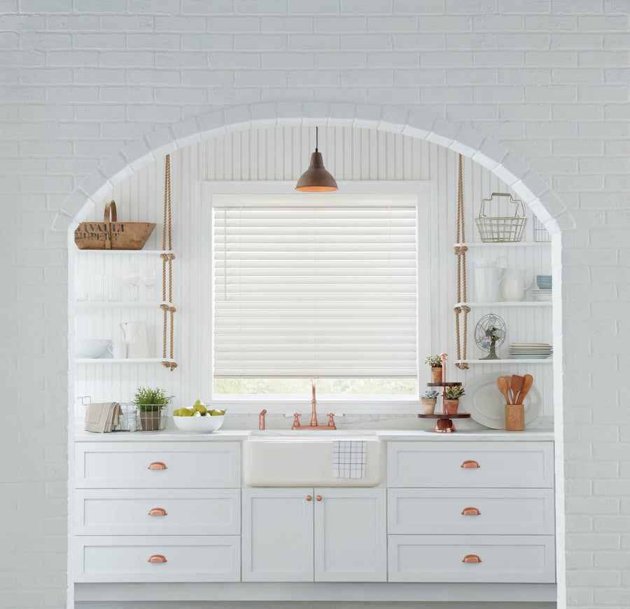 Bright white kitchen with a farmhouse sink, copper fixtures, open shelves, potted plants, and a window featuring Parkland® Wood Blinds beneath an arched brick opening.
