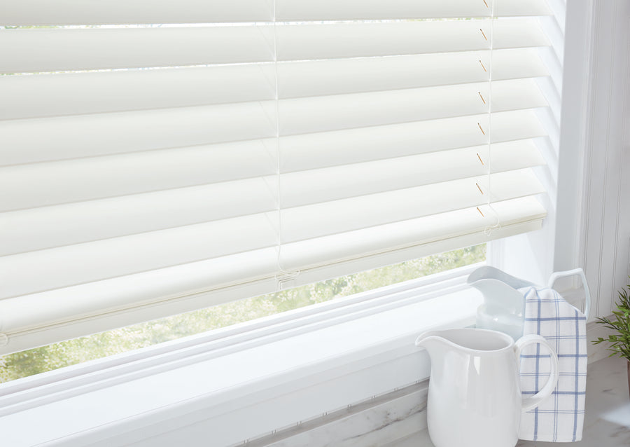 A close-up of a white window with partially closed Parkland® Wood Blinds. On the sill are a white ceramic pitcher, a white mug, a folded blue plaid cloth, and a small green plant as sunlight filters through the slats.