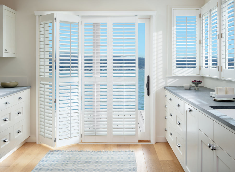 Bright kitchen with white cabinets and countertops, large sliding glass doors fitted with Palm Beach™ Polysatin™ Shutters. Sunlight streams in, revealing blue water and sky. A light rug accents the wooden floor.