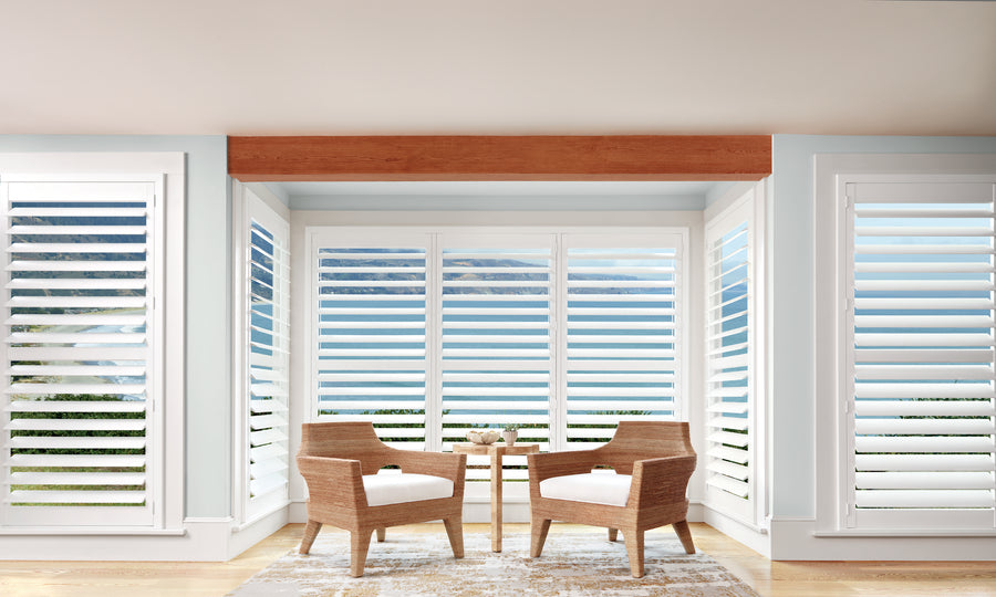Two wicker chairs with white cushions surround a small round table in a bright room with large windows featuring Palm Beach™ Polysatin™ Shutters, offering a scenic view. A light rug decorates the wooden floor.
