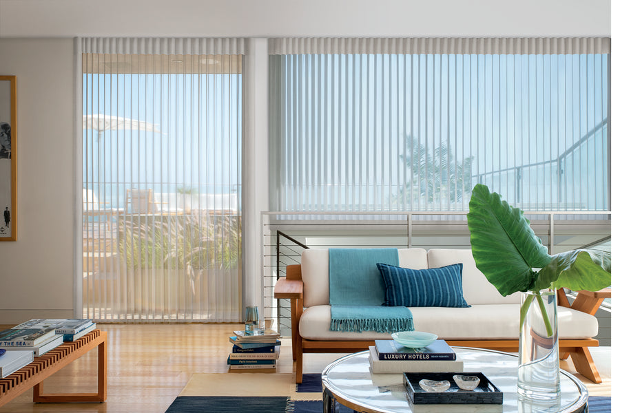 A modern living room with large windows dressed in Luminette® Privacy Sheers, allowing gentle natural light to filter in. The space includes a white sofa with blue cushions, a glass coffee table with books, and a vase holding a big green leaf.