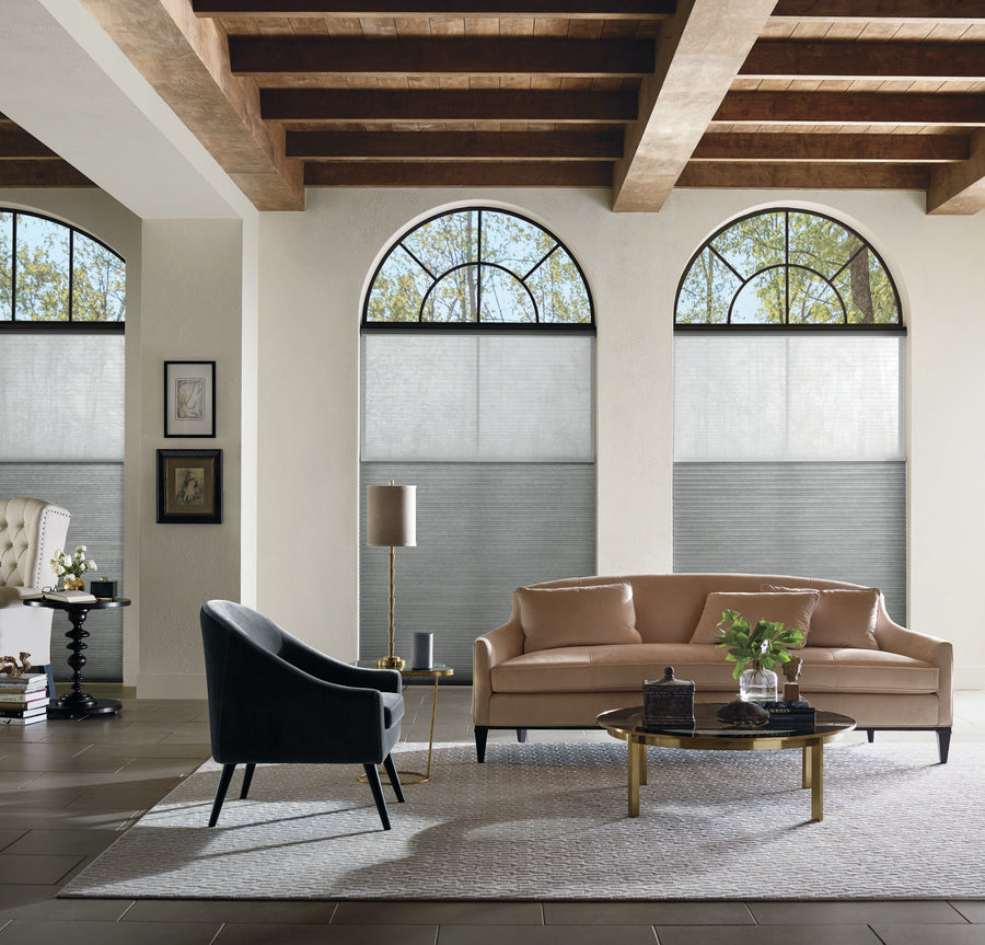 A modern living room features a tan leather sofa, black armchair, round coffee table, wooden ceiling beams, neutral decor on a patterned rug, and large arched windows adorned with Applause® Honeycomb Shades.