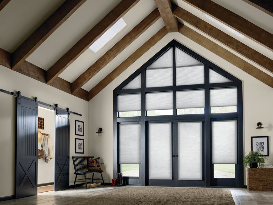 A modern living room with vaulted ceilings, exposed wooden beams, large black-framed windows featuring Applause® Honeycomb Shades, a sliding barn door, a bench with pillows, and a potted plant on the right.