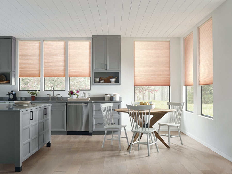 Bright, modern kitchen with gray cabinets, stainless steel appliances, a round wooden table with four white chairs, and large windows enhanced by Applause® Honeycomb Shades featuring double-cell construction for extra insulation.