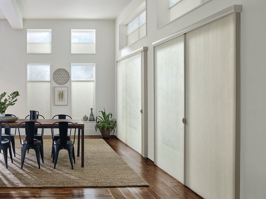 A bright dining room features a wooden table, black chairs, potted plants, wall art, and tall windows with Applause® Honeycomb Shades. Sliding doors with frosted glass cover one wall, and a woven rug lies on the hardwood floor.