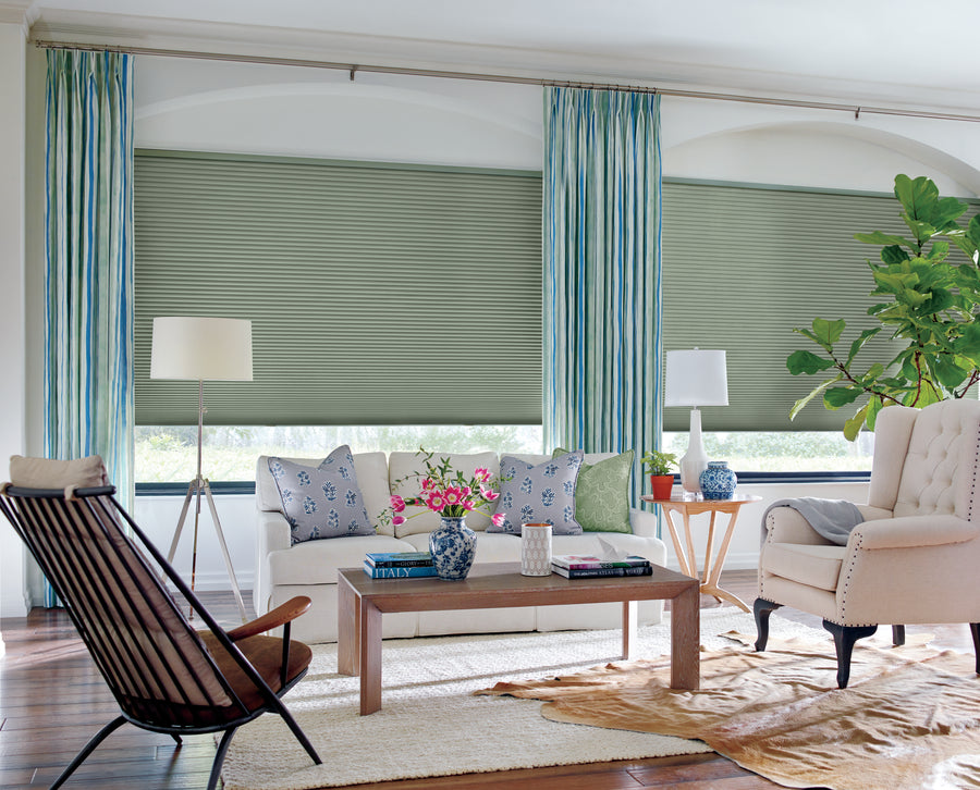 A bright living room with green Duette® Honeycomb Shades, blue-striped curtains, a white sofa with patterned pillows, a beige armchair, wooden chairs, potted plants, and a wooden coffee table decorated with flowers and vases.