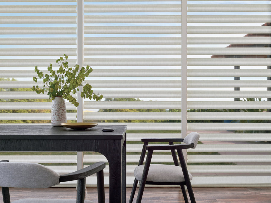 A modern dining area with a dark wood table, two cushioned chairs, a vase of greenery, and large windows outfitted with Pirouette® Window Shadings for stylish light control and UV protection.