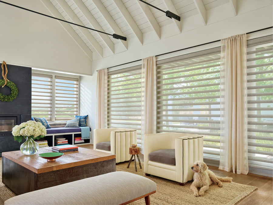 A bright, modern living room with large windows featuring Pirouette® Window Shadings for gentle light, sheer curtains, two cream armchairs, a wood coffee table with flowers and books, a beige bench, and a small dog resting on a woven rug.