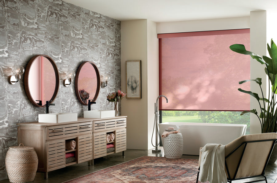 A modern bathroom features two round mirrors, twin sinks on a wooden vanity, a freestanding tub, patterned wallpaper, indoor plants, a decorative rug, and Designer Roller Shades in pink for premium window styling.