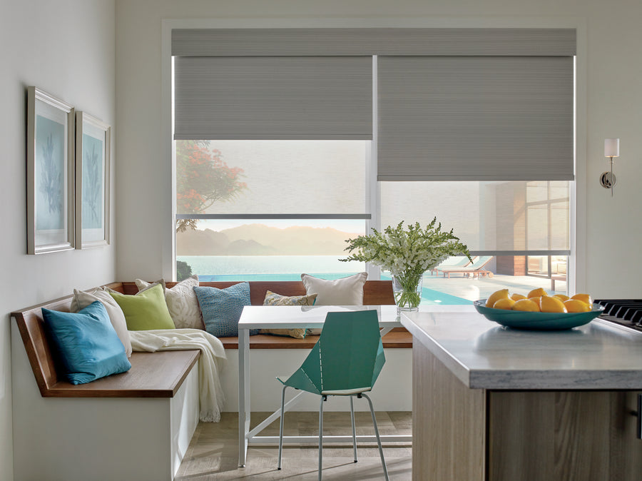 Bright, modern breakfast nook with a built-in wooden bench, colorful cushions, a white table, turquoise chair, flowers in a vase, and large windows featuring Designer Roller Shades overlooking a pool and scenic outdoor view.
