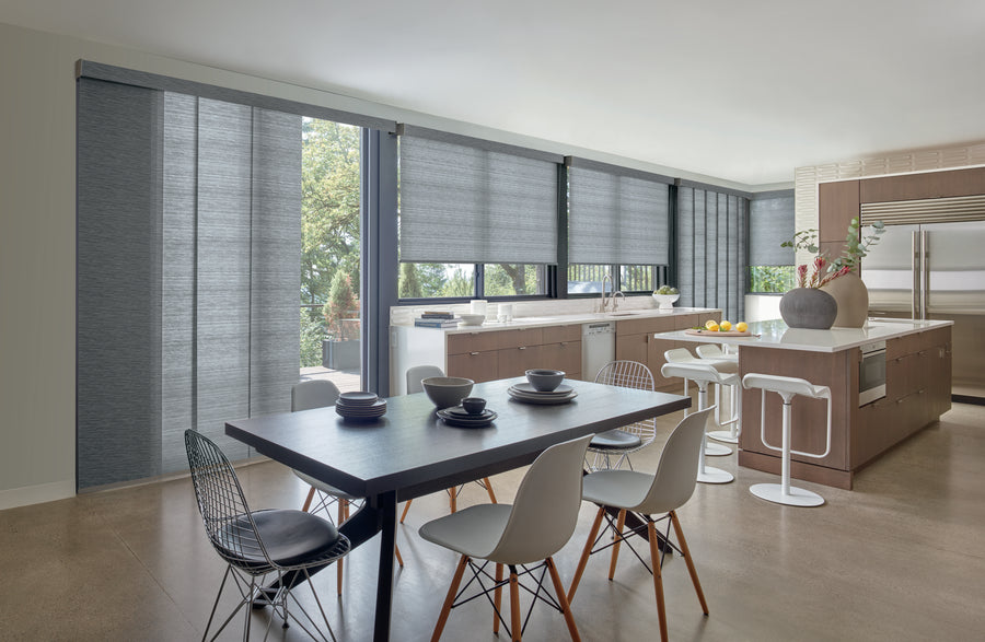 A modern kitchen and dining area with large windows features Skyline® Gliding Window Panels for a sleek look, a dark dining table seating six, an island with white barstools, and neutral decor bathed in natural light.