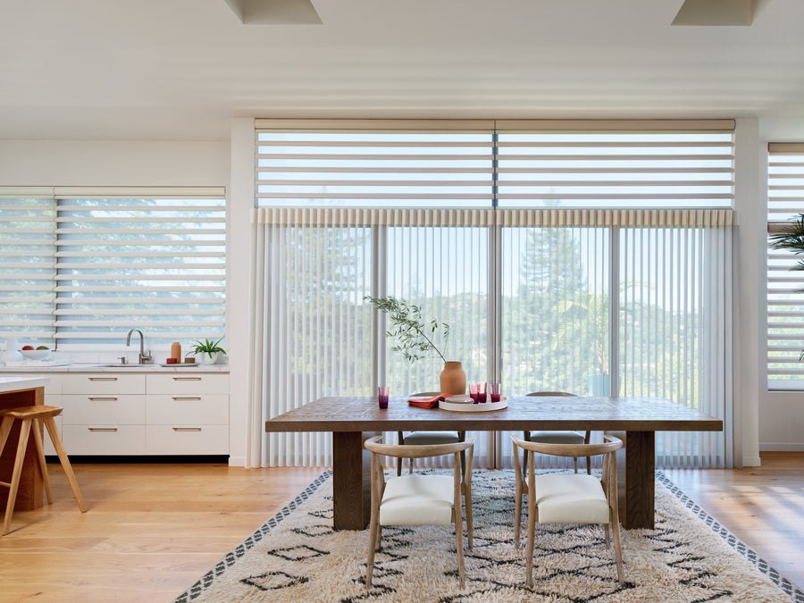 A modern dining room with large windows showcases Luminette® Privacy Sheers, a wooden table with decor, two chairs, a geometric rug, and views of trees outside. A white kitchen with stools appears to the left.