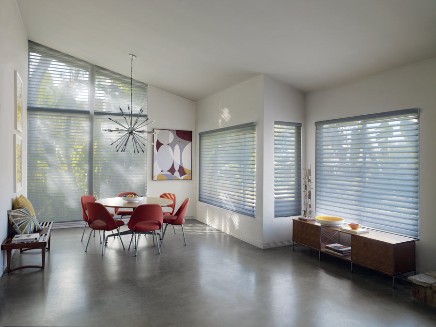 A bright, modern dining room features red chairs at a white table, contemporary chandelier, abstract art, and large windows with Nantucket™ Window Shadings for soft light diffusion. A sideboard with decorative bowls sits on polished concrete floors.