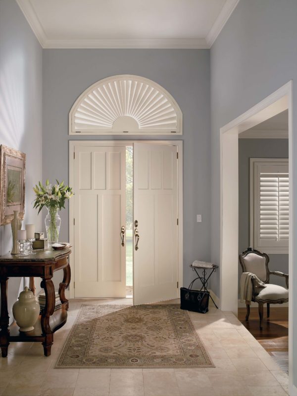 A bright entryway with double white doors topped by a half-circle window featuring affordable NewStyle® Hybrid Shutters, a patterned rug, wooden console table with flowers, and a classic armchair in the adjacent room.
