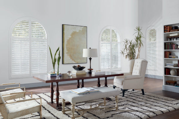 A bright, elegant home office with arched windows features affordable NewStyle® Hybrid Shutters, a wooden desk, upholstered chairs, a lamp, potted plants, a large rug, wall art, and a built-in bookshelf filled with books and decor.