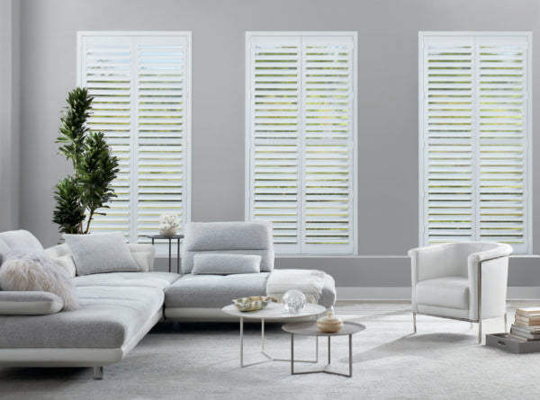 Modern living room with a light gray sectional, white armchair, small round coffee tables, and three tall windows featuring affordable NewStyle® Hybrid Shutters that let in natural light. Neutral, minimalist décor and a touch of green from a houseplant.