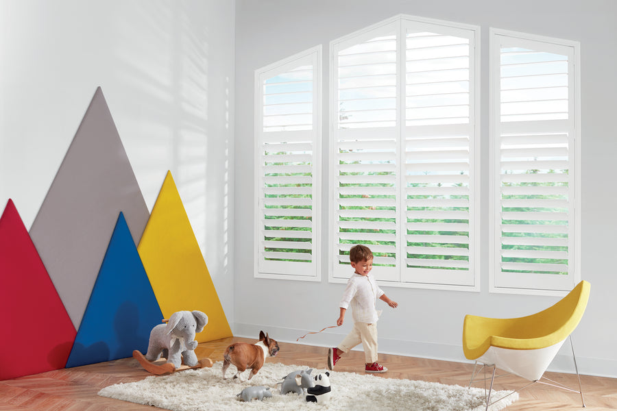 A young child walks a toy dog on a leash in a bright, modern room with NewStyle® Hybrid Shutters, a yellow chair, plush toys, and colorful geometric mountain-shaped wall art.