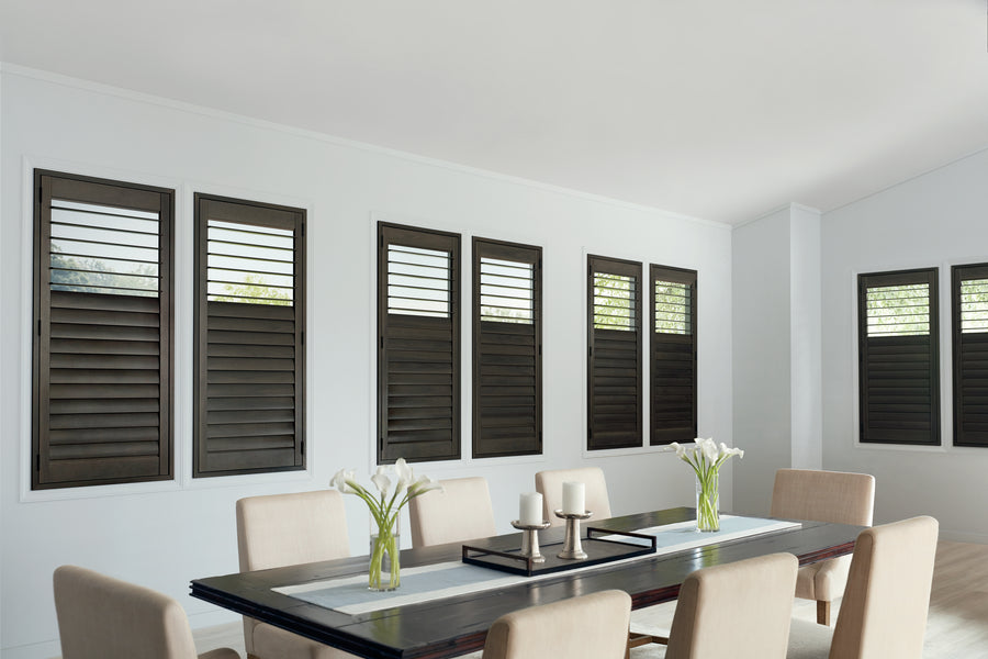 Modern dining room with beige chairs and a dark wood table adorned with white calla lilies and candles. The windows on the crisp white wall are covered with NewStyle® Hybrid Shutters in a wood-look finish.