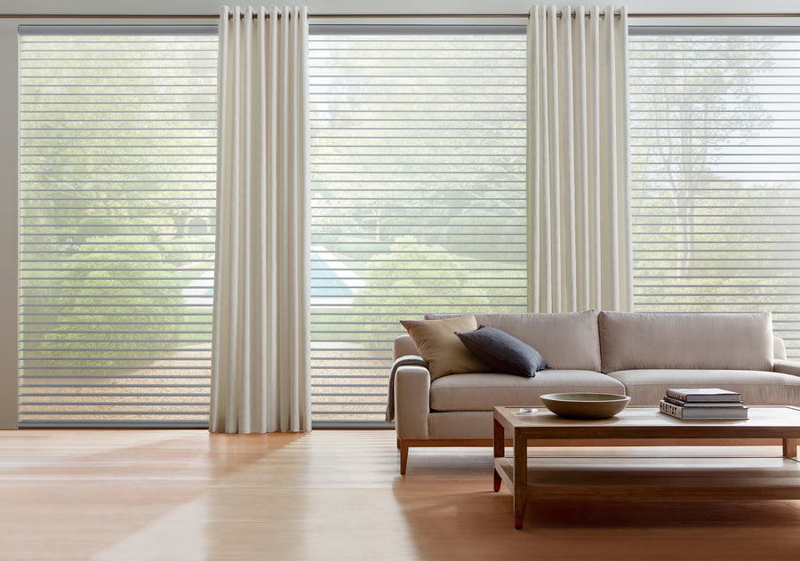 A modern living room features Nantucket™ Window Shadings, gently diffusing light through sheer blinds and white curtains. A beige sofa, wooden coffee table, and decor rest on a light wood floor, with trees and a house seen outside large windows.