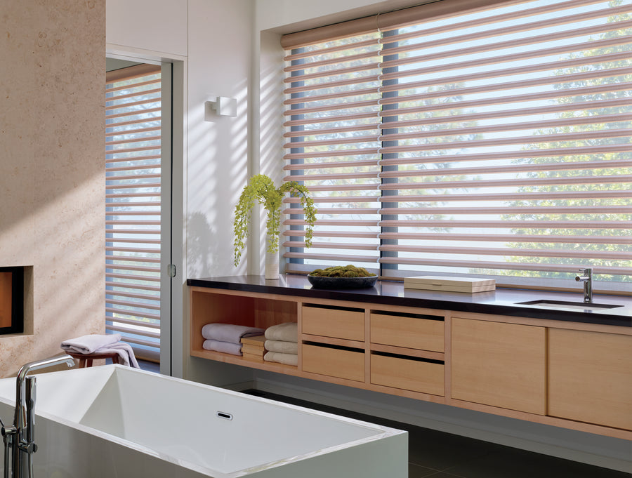 Modern bathroom with large bathtub, wooden cabinets, stacked towels, a vase with green flowers, and a wide window fitted with Pirouette® Window Shadings for elegant light control and UV protection.