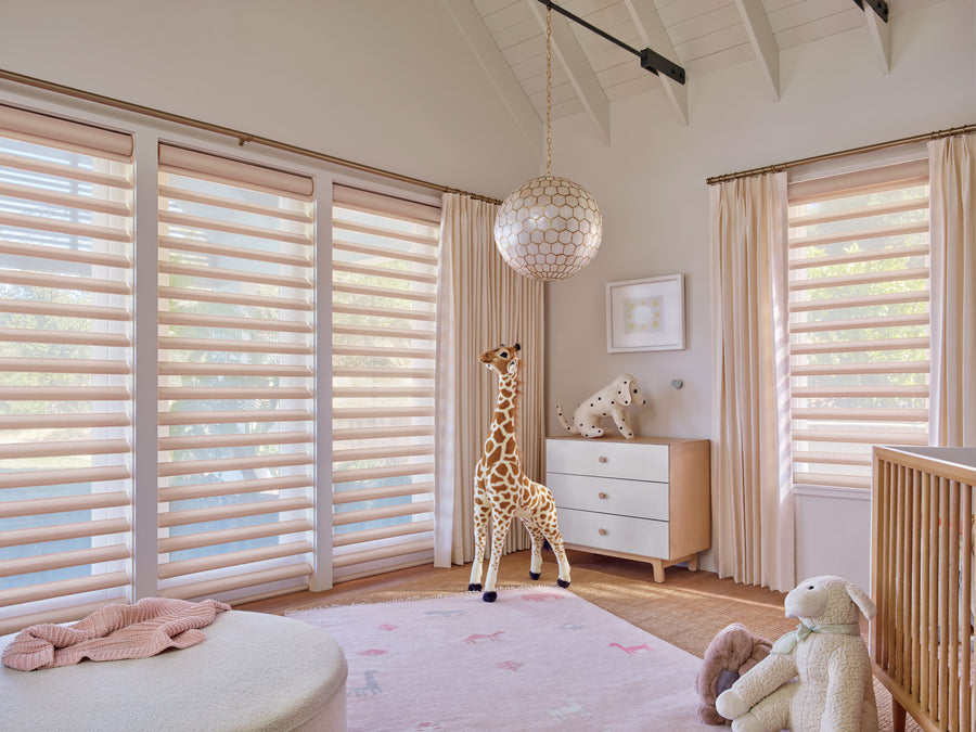 Bright nursery with large windows, Pirouette® Window Shadings for UV protection and light control, a plush giraffe, dresser with dalmatian toy, crib, pale curtains, round ottoman, and a soft pink animal-themed rug.
