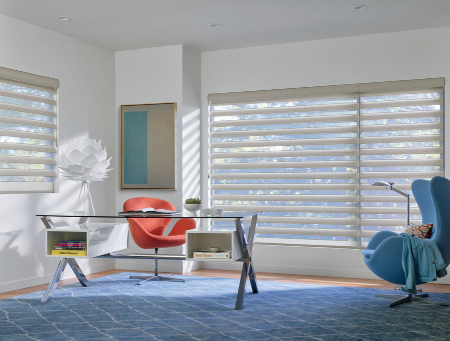 Modern home office with large windows dressed in Pirouette® Window Shadings for soft light and UV protection, a glass desk, orange and blue armchairs, geometric rug, sculptural white lamp, potted plant, and sunlight streaming in.