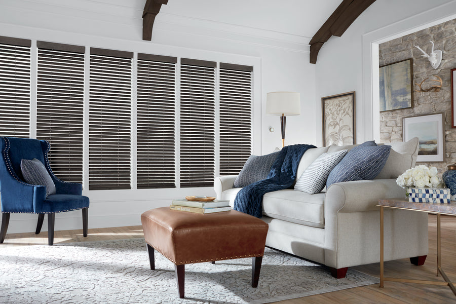 A cozy living room with a light sofa, blue accent chair, brown leather ottoman, floor lamp, pillows, wall art, and large windows adorned with Parkland® Wood Blinds featuring rich hardwood slats for natural light.