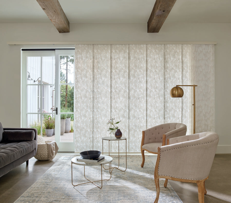 A modern living room with a gray sofa, beige armchairs, round coffee table, gold floor lamp, Skyline® Gliding Window Panels as window treatment, a rug, exposed wooden beams, and glass doors opening to an outdoor patio.