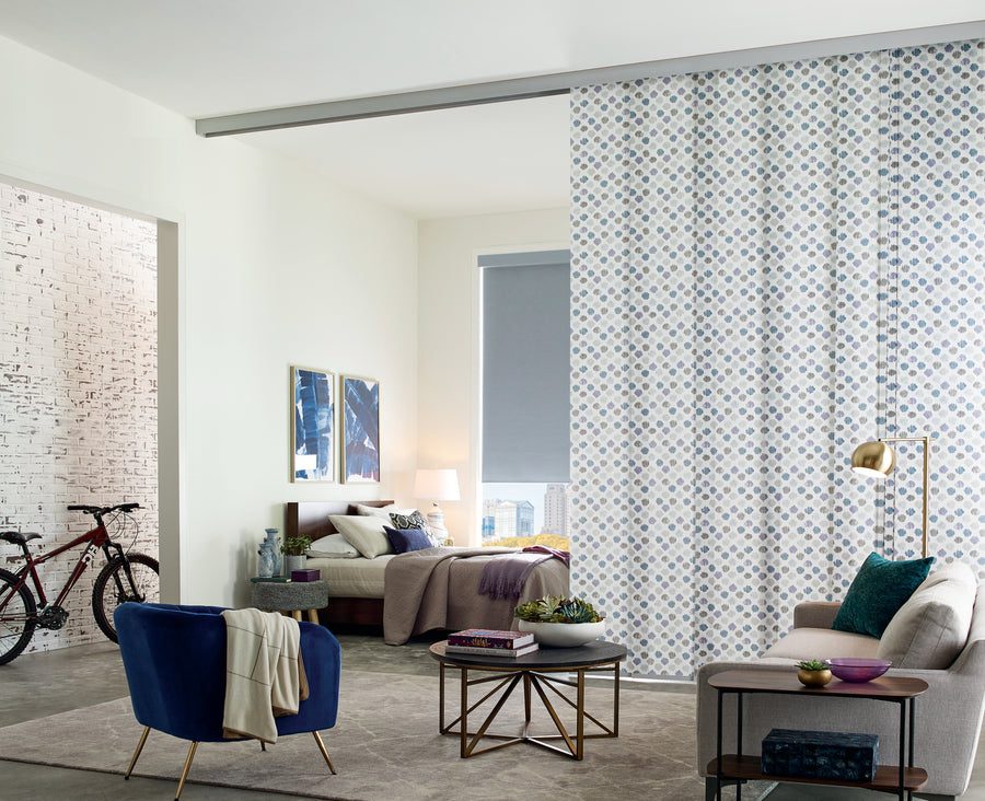 Modern living room featuring a blue armchair, beige sofa, round coffee tables, and Skyline® Gliding Window Panels as a divider; a bicycle and two paintings appear by the white brick wall in the background.
