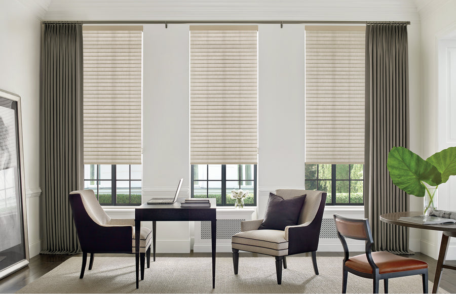 A modern office features a dark desk, two upholstered chairs, a laptop, and large windows with Sonnette™ Cellular Roller Shades in beige and gray curtains. A smaller table with a plant and chair sits nearby in this bright, minimalistic space.