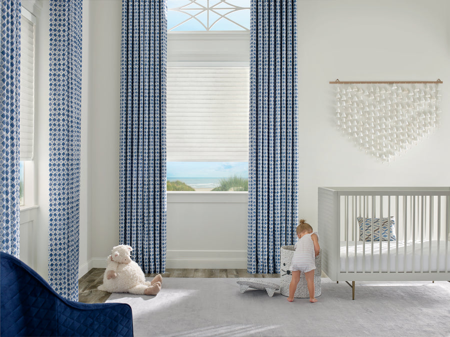 A nursery featuring Sonnette™ Cellular Roller Shades, tall blue patterned curtains, a white crib, plush sheep, and a baby in white by a pillow. Sunlight filters in through the window, showcasing a scenic outdoor view.