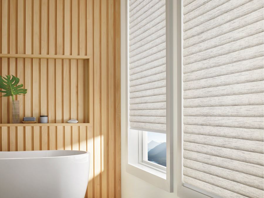 A modern bathroom with light wood slat walls, a white freestanding tub, decor on a small shelf, and a window featuring Sonnette™ Cellular Roller Shades partially covering scenic mountain views.