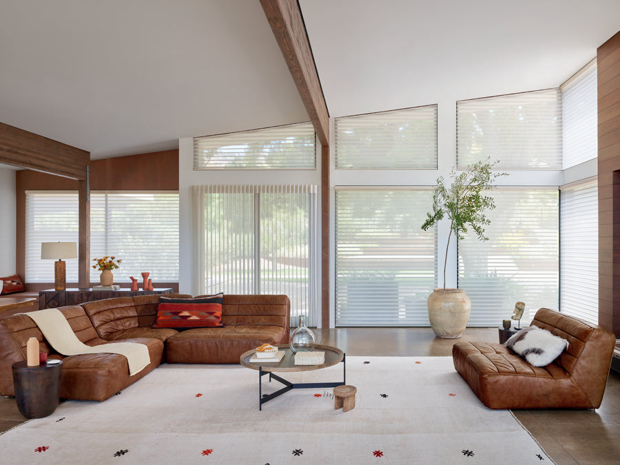 A modern living room with large windows dressed in Luminette® Privacy Sheers, a brown leather sectional and chair, a round coffee table, a white patterned rug, and a tall vase with branches; natural light fills the space.