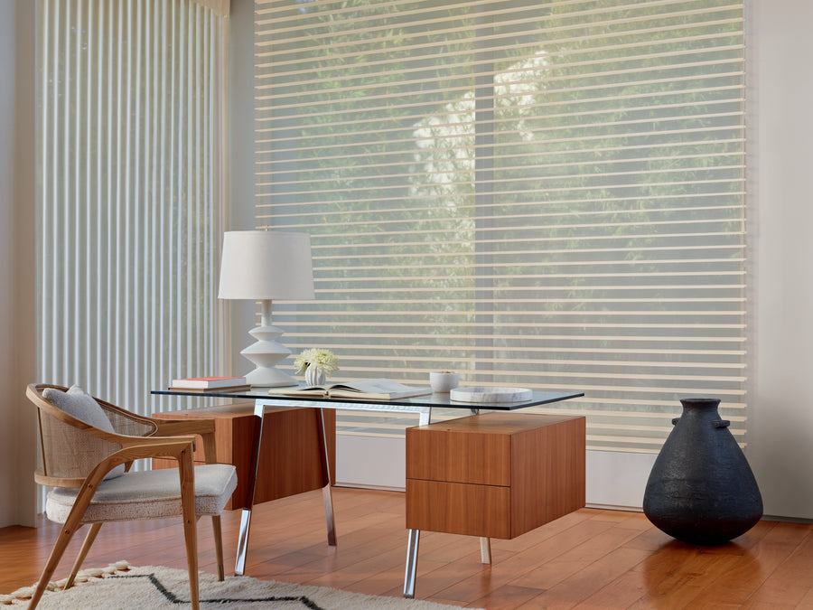 A modern home office with a glass desk, wooden drawers, a white lamp, an open book, mug, wicker chair, dark vase, and floor-to-ceiling Luminette® Privacy Sheers gently filtering natural light.