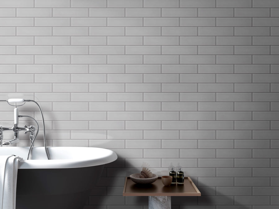 A white bathtub with a towel sits beside a small wooden table holding a brush, bowl, and bottles, set against a wall of Luna 3x12 Glass Subway Tile in light gray.