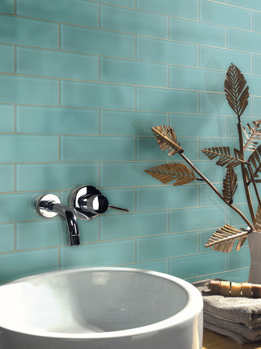 A modern bathroom sink with a chrome faucet is set against a wall of Luna 3x12 Glass Subway Tile in glossy turquoise. Decorative metallic leaves and folded towels sit neatly beside the basin.