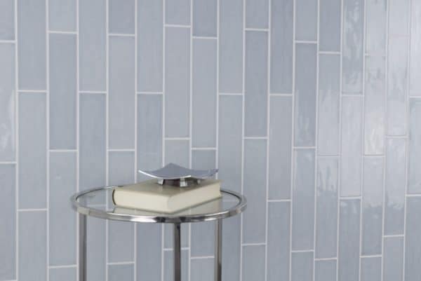 A round metal table with a white book and silver décor sits in front of a wall covered in glossy, light blue Habitat 2.5x10.3 Subway Tile arranged in a vertical brick pattern.