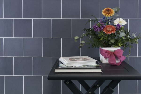A small black table stands against a Habitat 5.1x5.1 Subway Tile blue backsplash, displaying a stack of books and a flower arrangement in a white pot with a bright pink ribbon.
