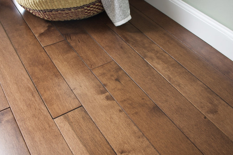 Close-up of a clean, shiny Acadia 3 1/4" Hard Maple Hardwood floor in medium brown. A woven basket with draped fabric appears in the upper left, and a white baseboard lines the wall.