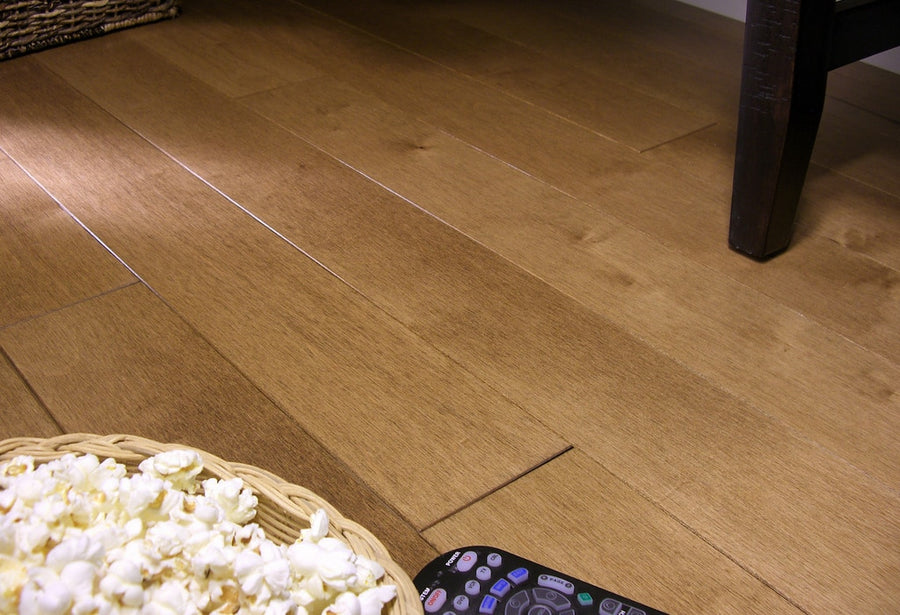 A close-up of Acadia 3 1/4" Hard Maple Hardwood floors with a basket of popcorn and a remote control near a dark wooden table leg.