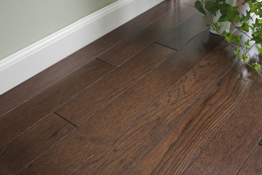 Dark brown Acadia 3 1/4" Hickory Hardwood flooring with rich grain patterns contrasts with a light baseboard and wall, while a potted green plant in the corner highlights its timeless, traditional style.