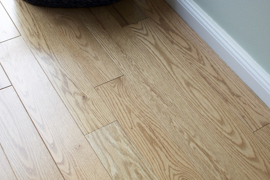 Acadia 3 1/4" Red Oak Hardwood flooring in light brown with natural grain patterns is shown next to a white baseboard and pale green wall. A woven basket adds warmth to these durable, traditional-style floors.