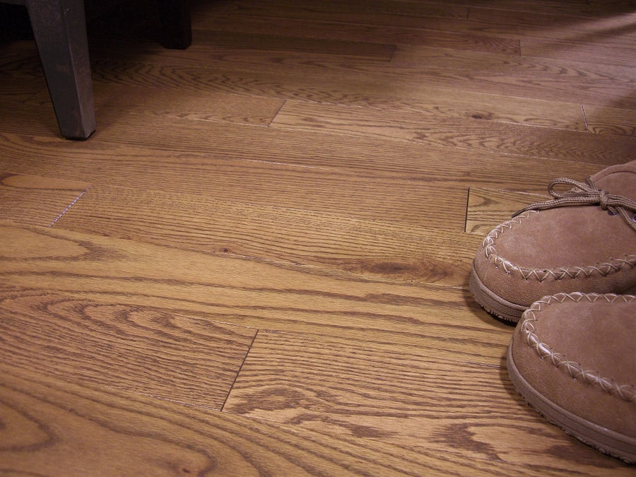 Close-up of Acadia 3 1/4" Red Oak Hardwood flooring, featuring light brown slippers on the right and a dark wooden chair leg in the upper left corner.