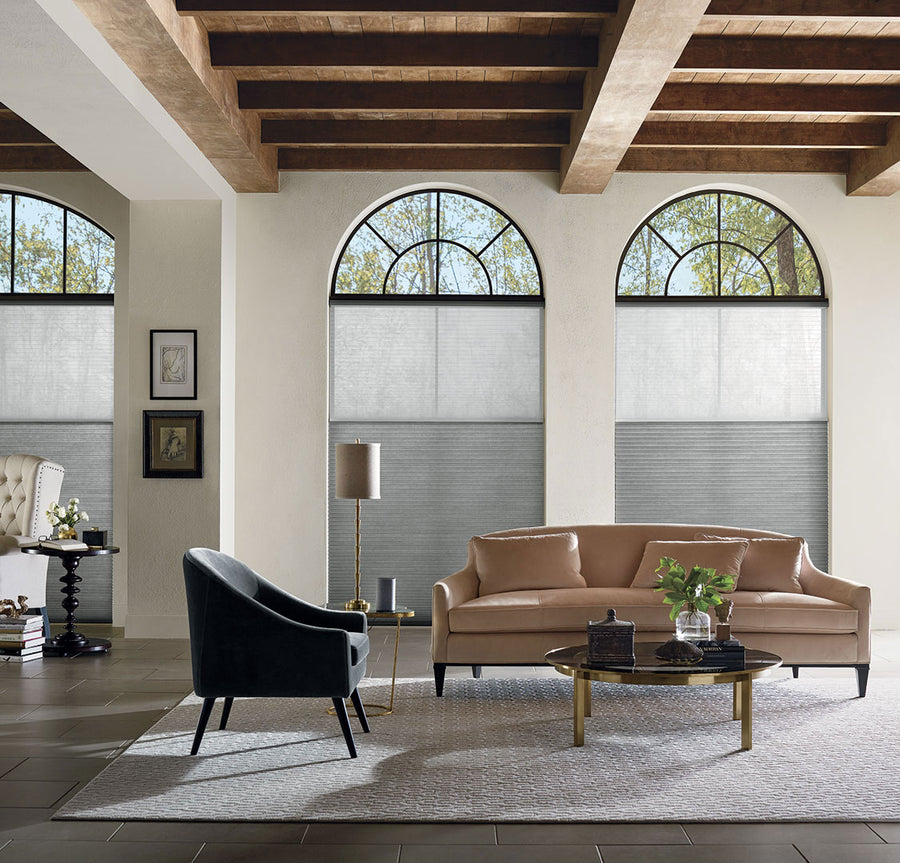 A stylish living room features a tan sofa, black armchair, glass coffee table, wooden ceiling beams, neutral decor, and large arched windows with Applause® Honeycomb Shades with PowerView® Motorization for energy efficiency.